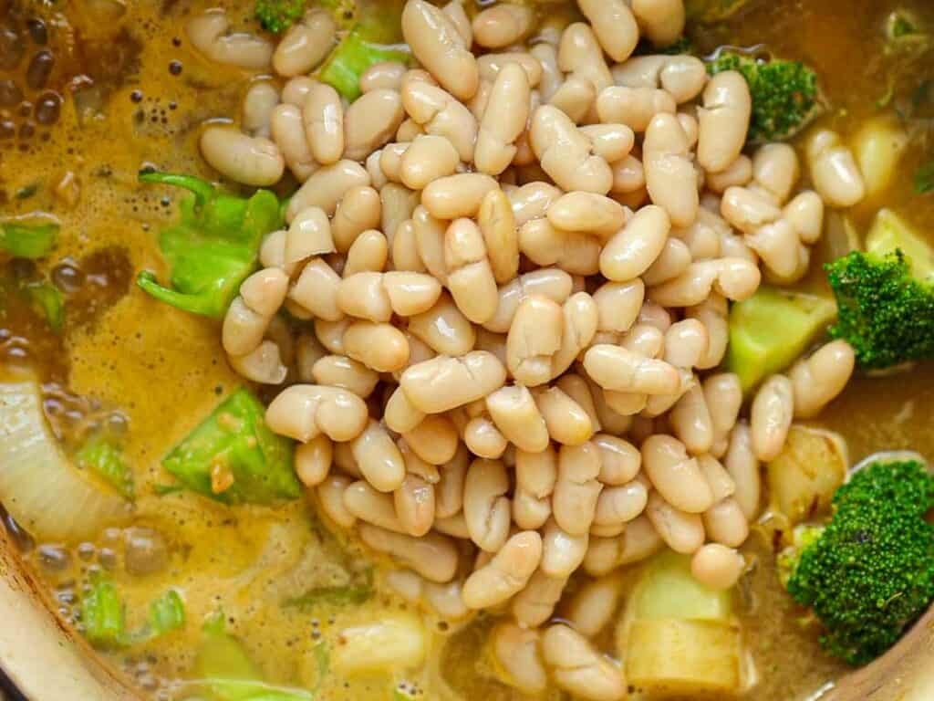 A pot of soup with white beans, broccoli florets, and chopped onions in a broth. The ingredients are mixed and simmering, creating a hearty and colorful dish.